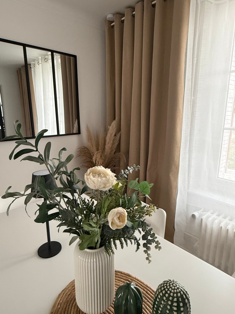 Un vase avec des fleurs artificielles sur une table, entouré de décorations modernes et de rideaux beiges.